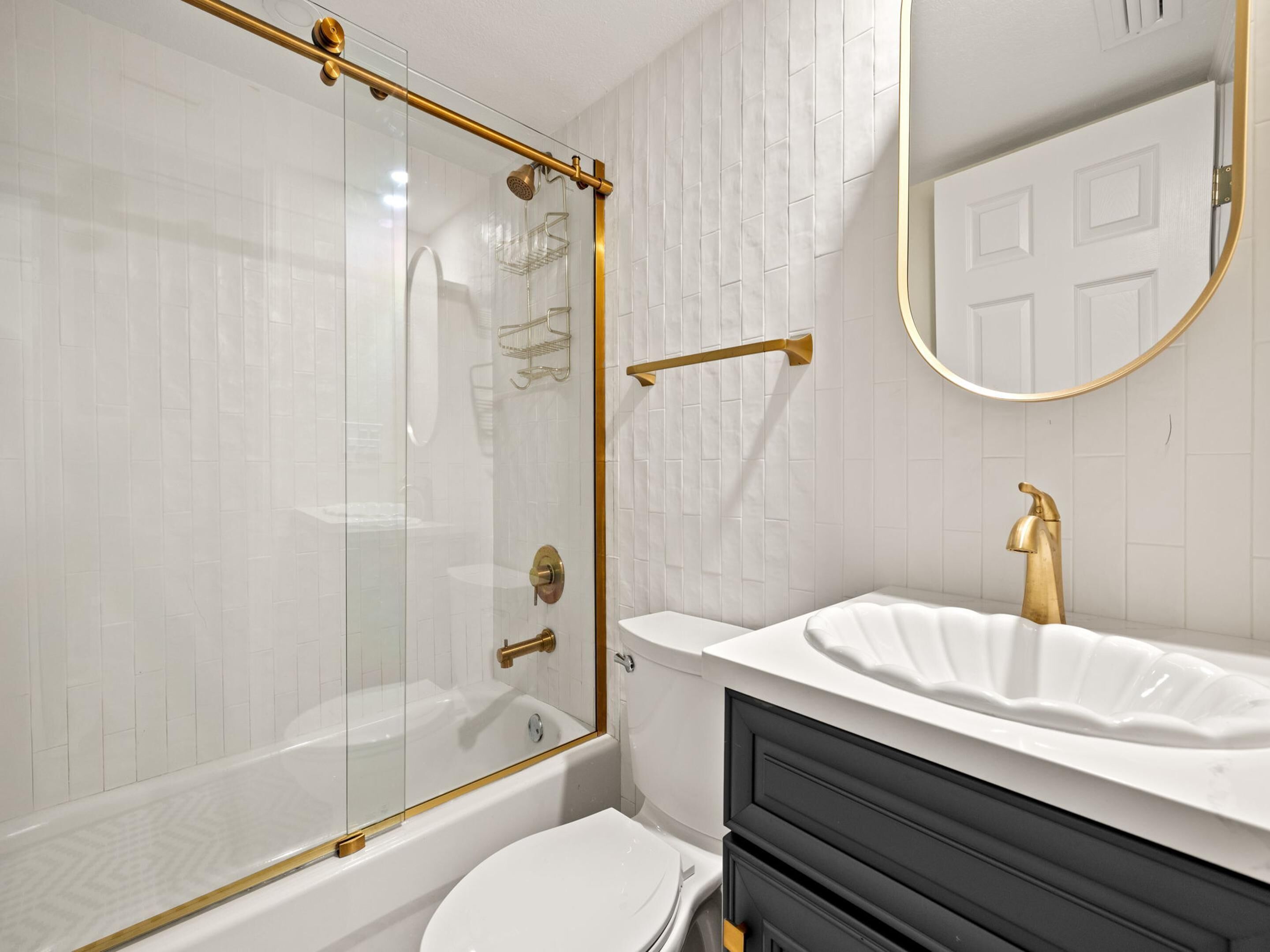 A modern bathroom with a glass-enclosed shower, a white bathtub, a white sink with a gold faucet, and a black vanity.