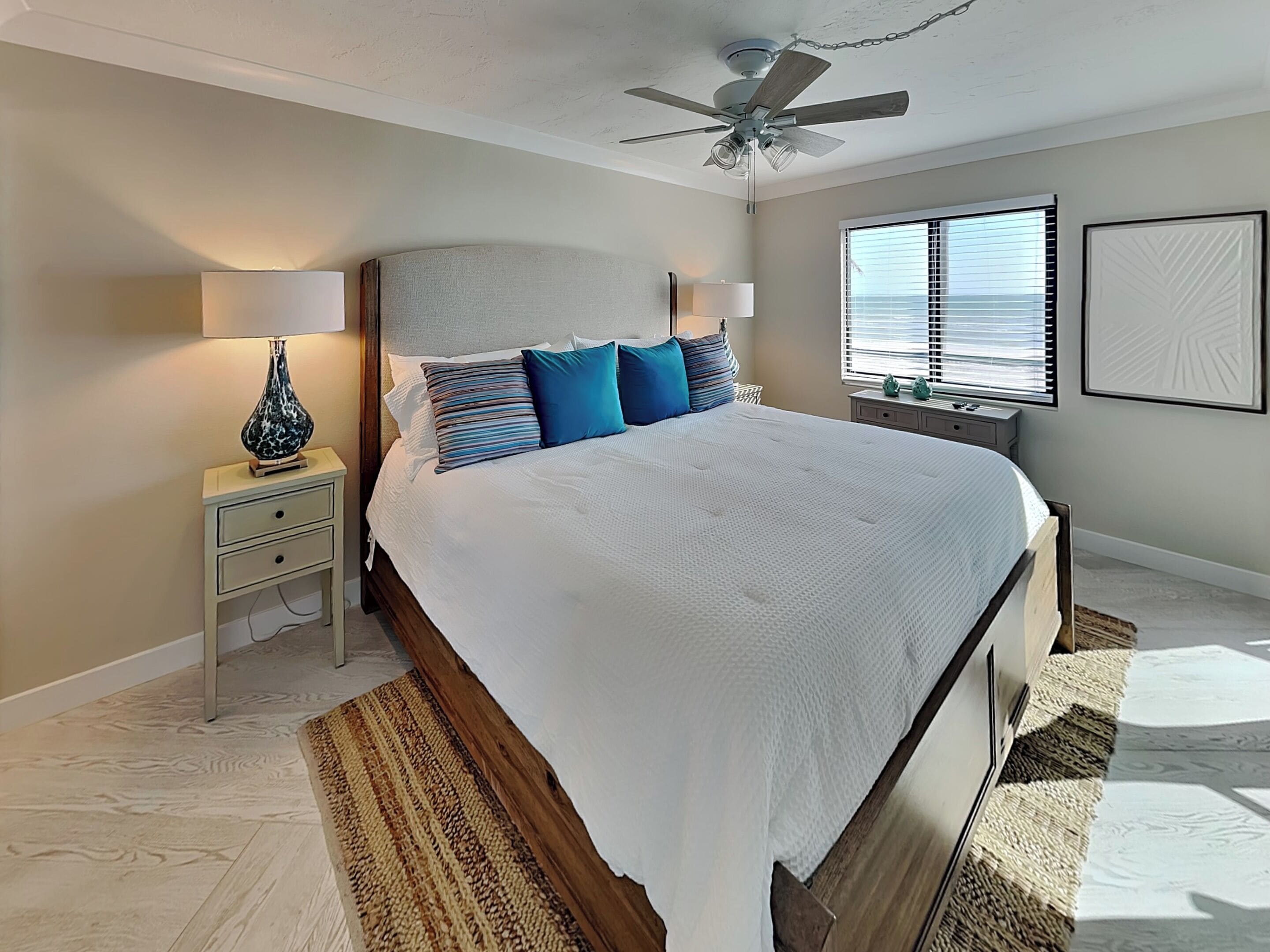 A bedroom with a large bed, a nightstand with a lamp, a ceiling fan, a window with blinds, and a wall-mounted artwork.