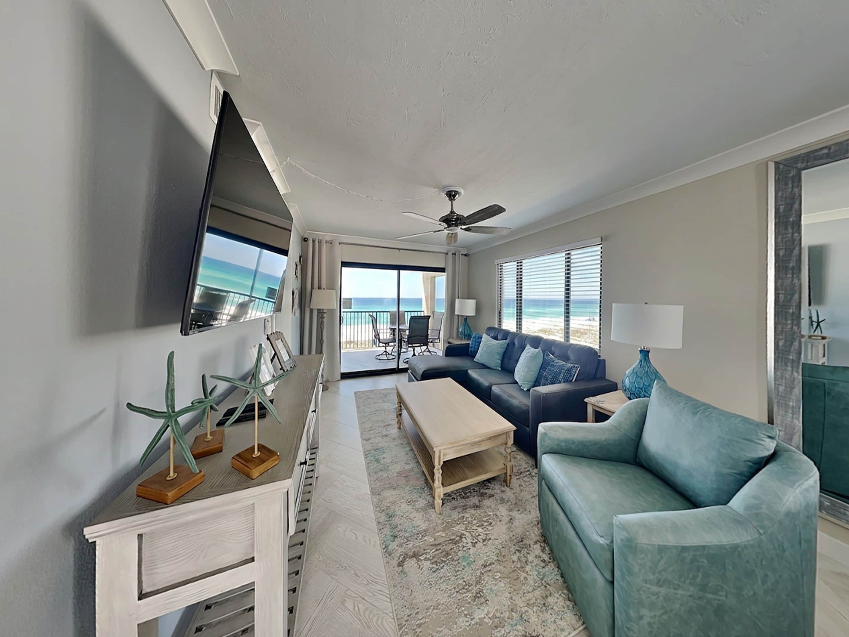 A modern living room with a large TV, a sofa, a coffee table, and a view of the beach through the glass door.