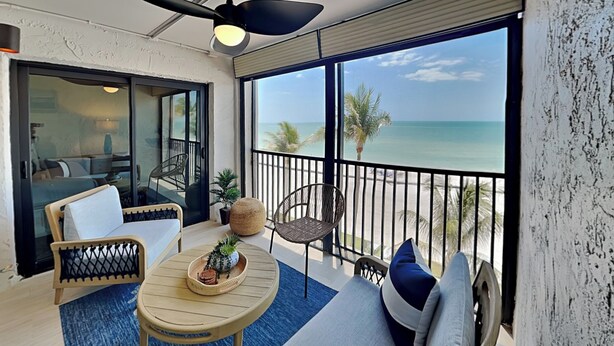 A balcony with a ceiling fan, a sofa, a small table with decorative items, and a view of the ocean. - Seaside 3 by Distinctive Beach Rentals (Fort Myers Beach)