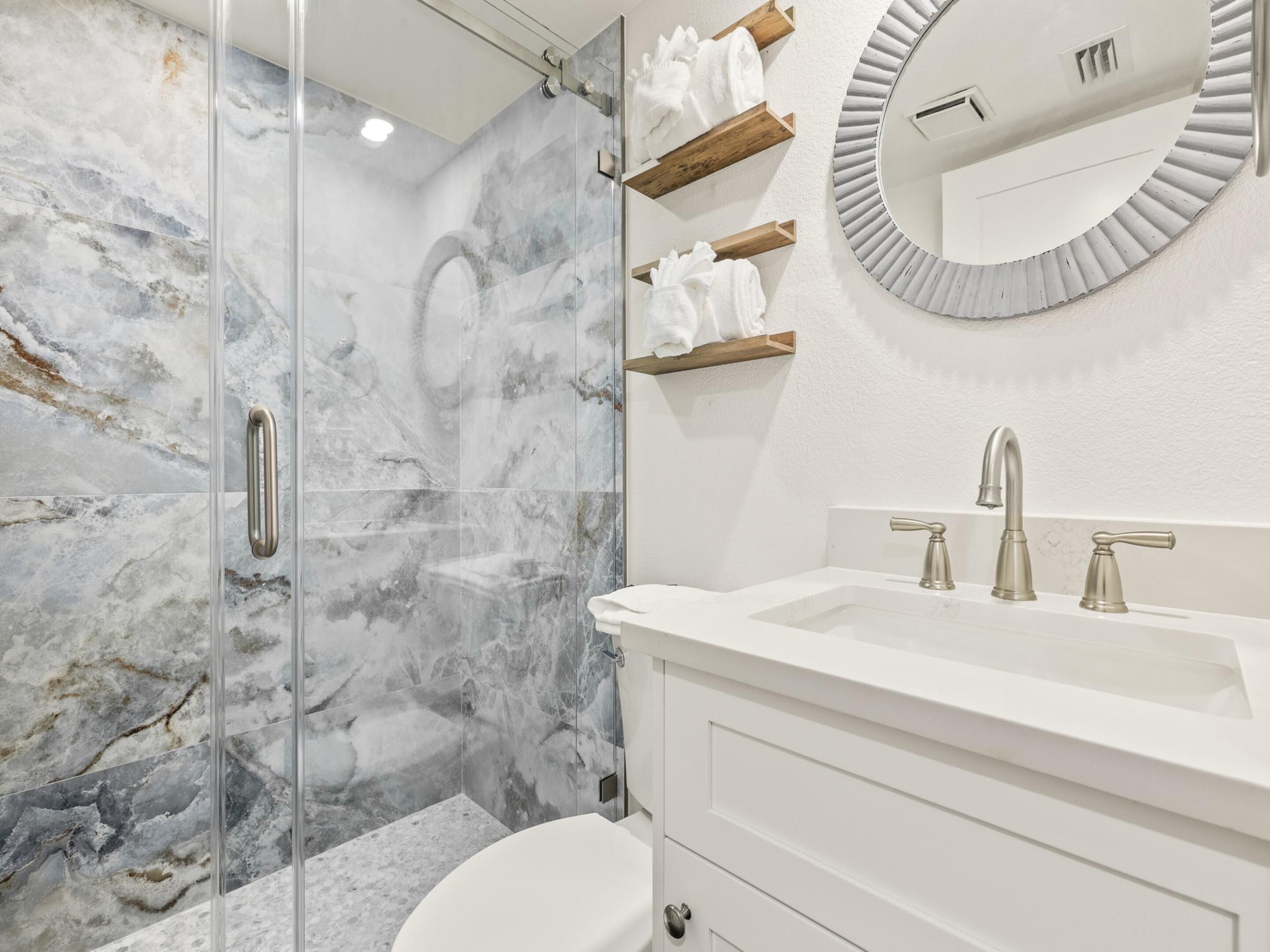 A modern bathroom with a glass shower enclosure, a white sink with a single faucet, and a round mirror with a distressed frame.