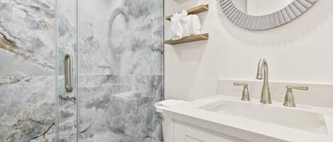 A modern bathroom with a glass shower enclosure, a white sink with a single faucet, and a round mirror with a distressed frame.