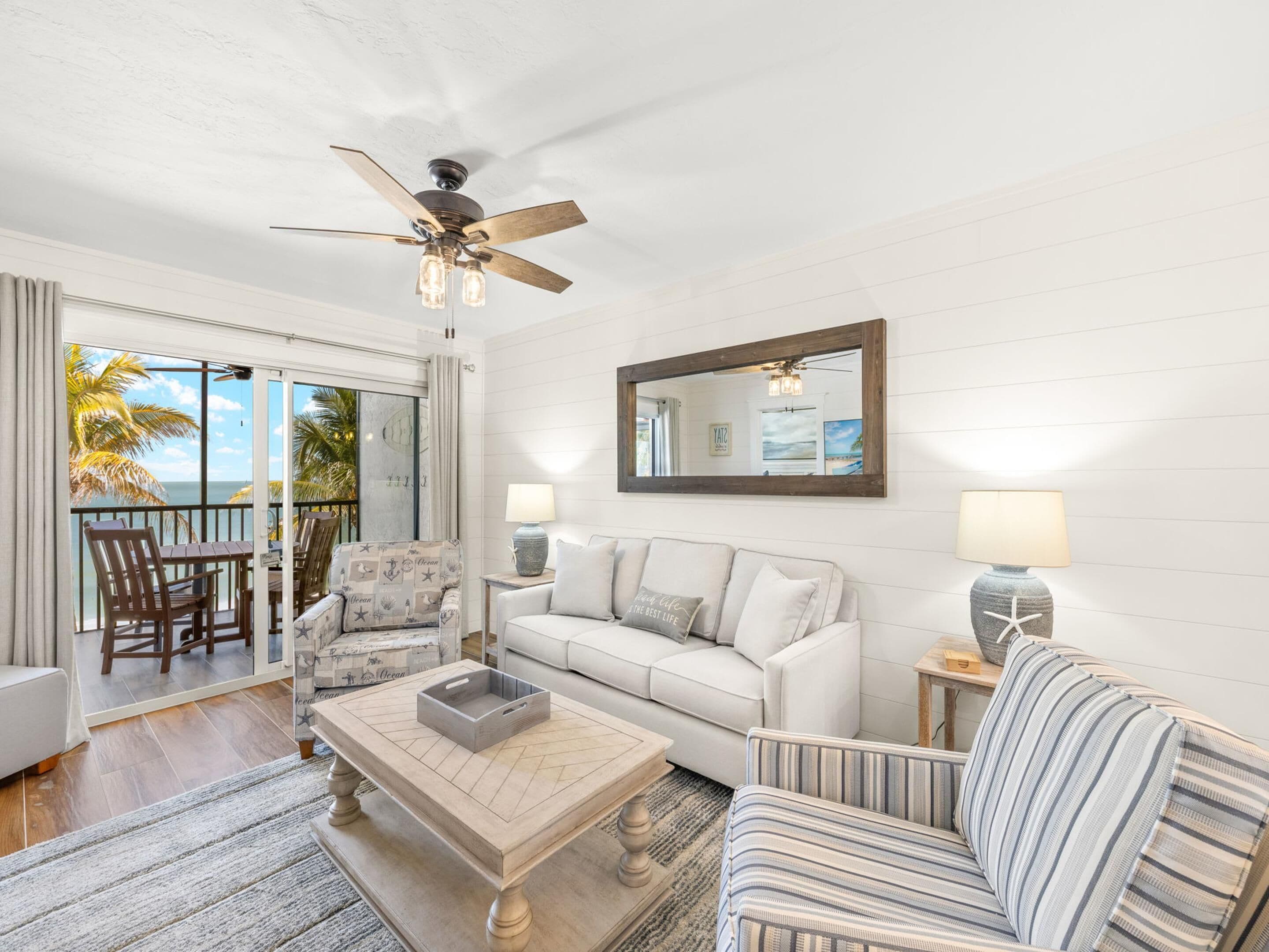 A spacious living room with a view of the ocean, a ceiling fan, a sofa, a striped armchair, a coffee table, and a side table with a lamp.