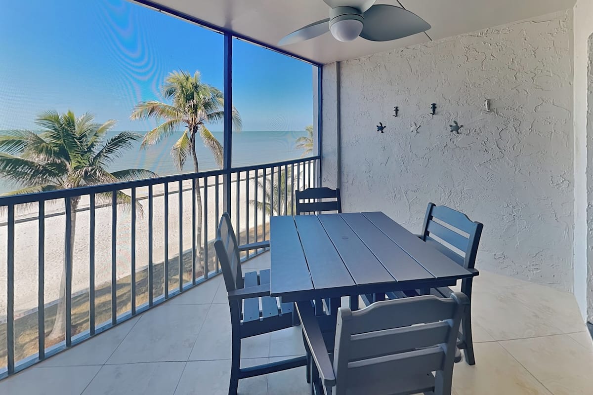 Uma varanda com mesa e cadeiras, com vista para uma praia com palmeiras.