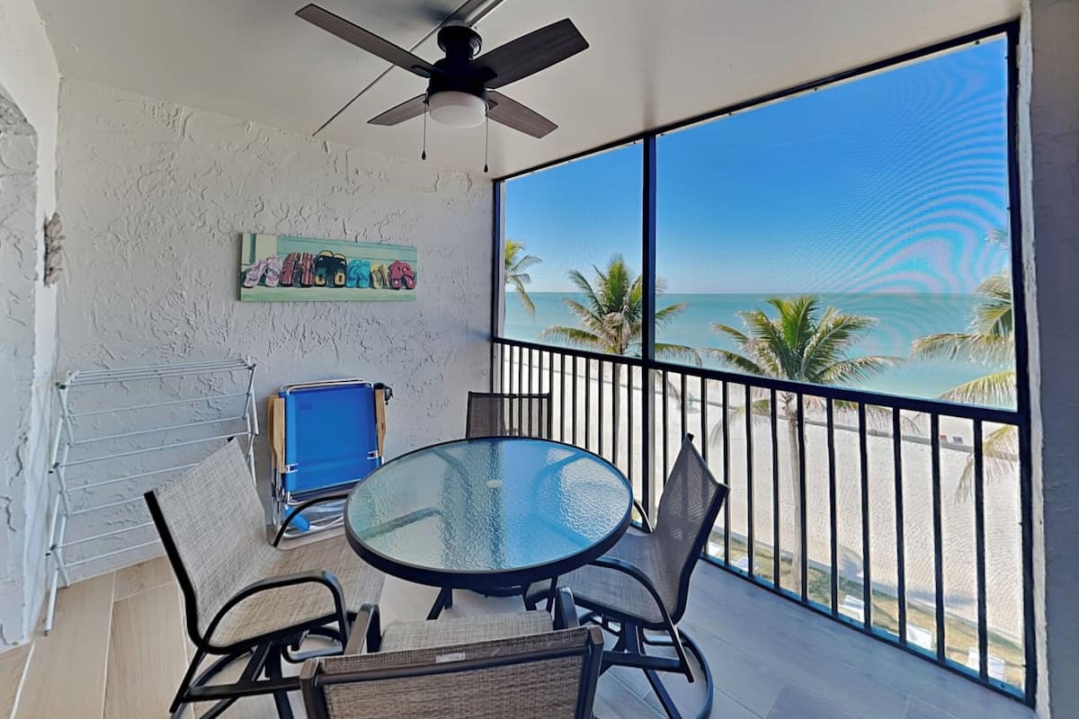 Uma varanda com mesa e cadeiras de vidro, um ventilador de teto, uma pequena estante com livros e vista para a praia e o oceano.