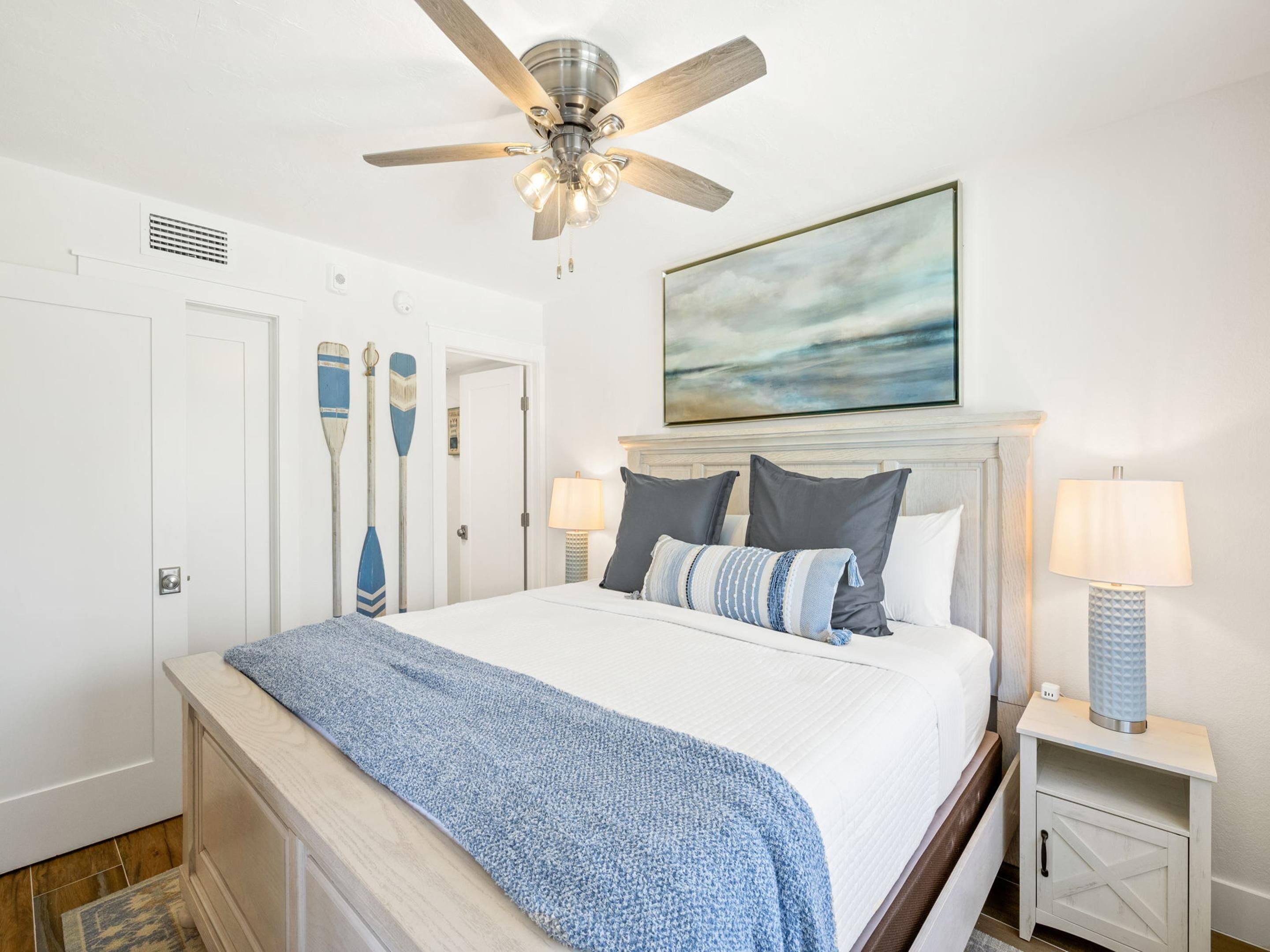 A bedroom with a bed, a ceiling fan, a painting, and a nightstand with a lamp.