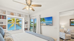 A bedroom with a bed, a ceiling fan, a bookshelf, a TV, and a view of the beach through a sliding door. - Seaside 3 by Distinctive Beach Rentals (Fort Myers Beach)