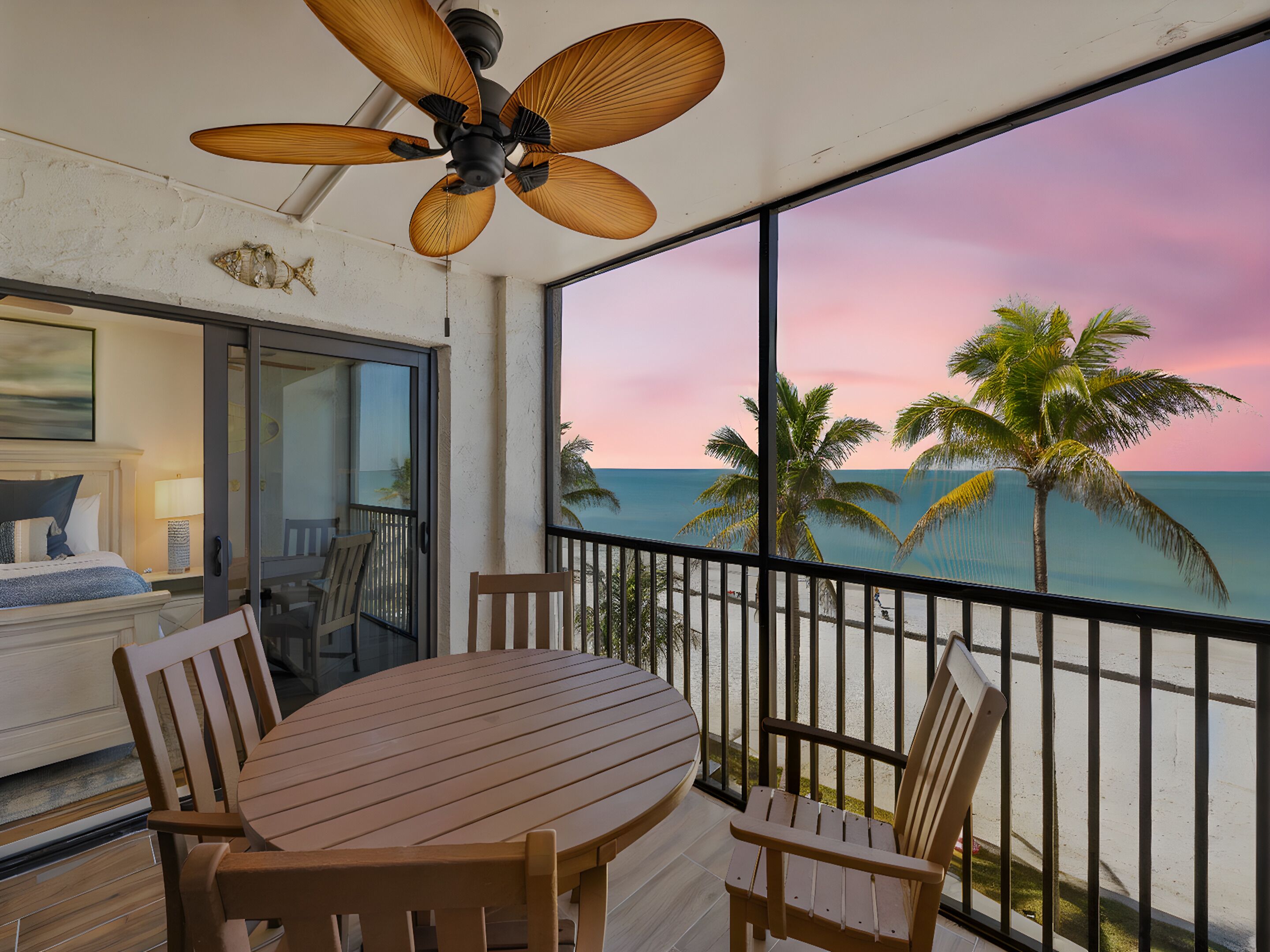 Un balcon avec une table et des chaises en bois, un ventilateur de plafond, et une vue sur l’océan et les palmiers au coucher du soleil.