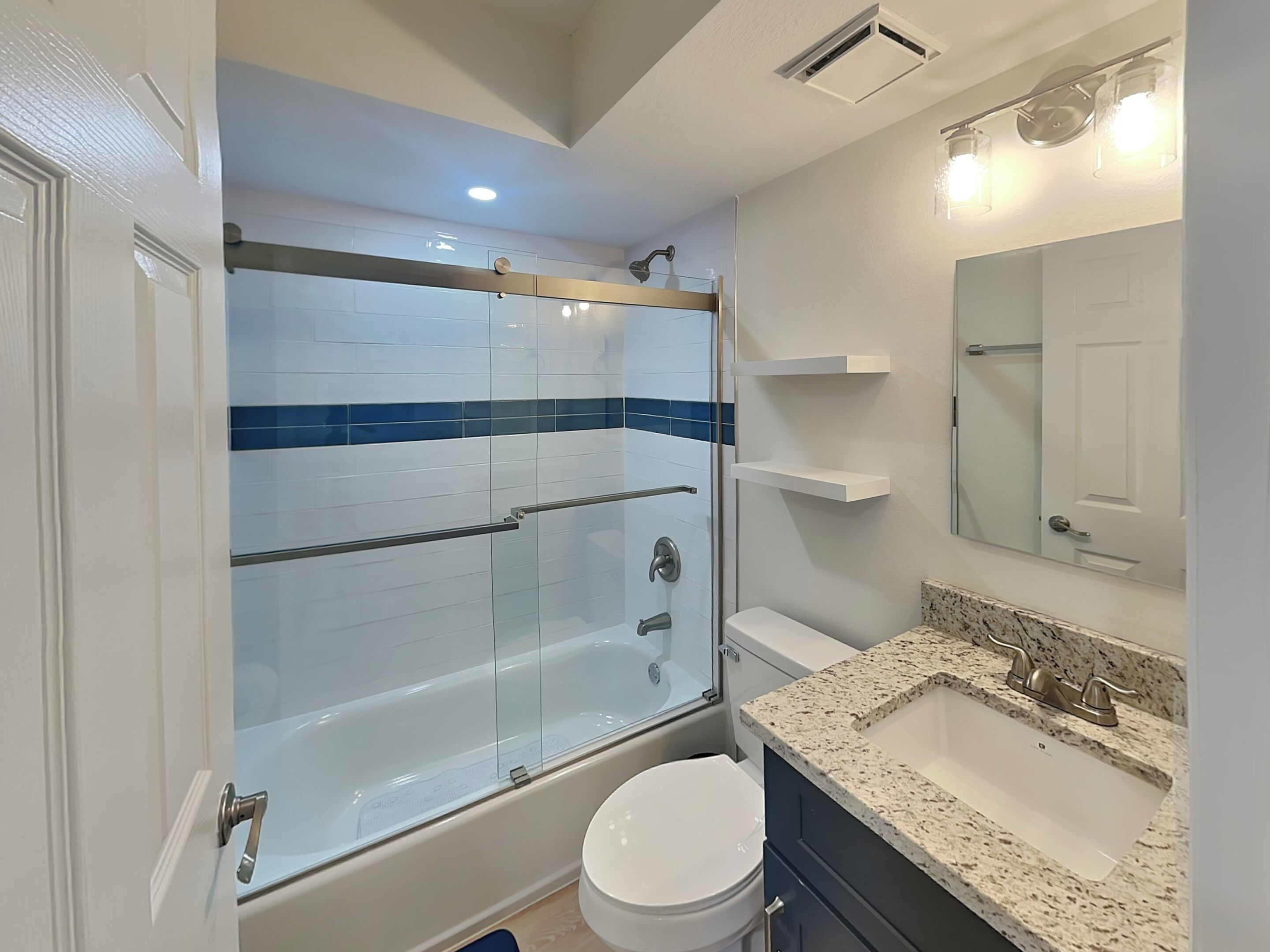 Un baño moderno con ducha envuelta en vidrio, inodoro blanco, lavamanos con encimera de granito y espejo sobre el lavamanos.