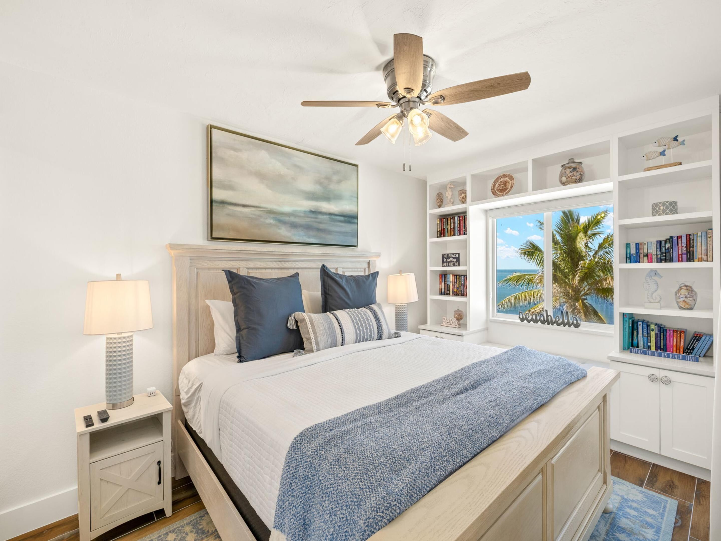 A bedroom with a bed, nightstands, a ceiling fan, a bookshelf, and a window with a view of palm trees.