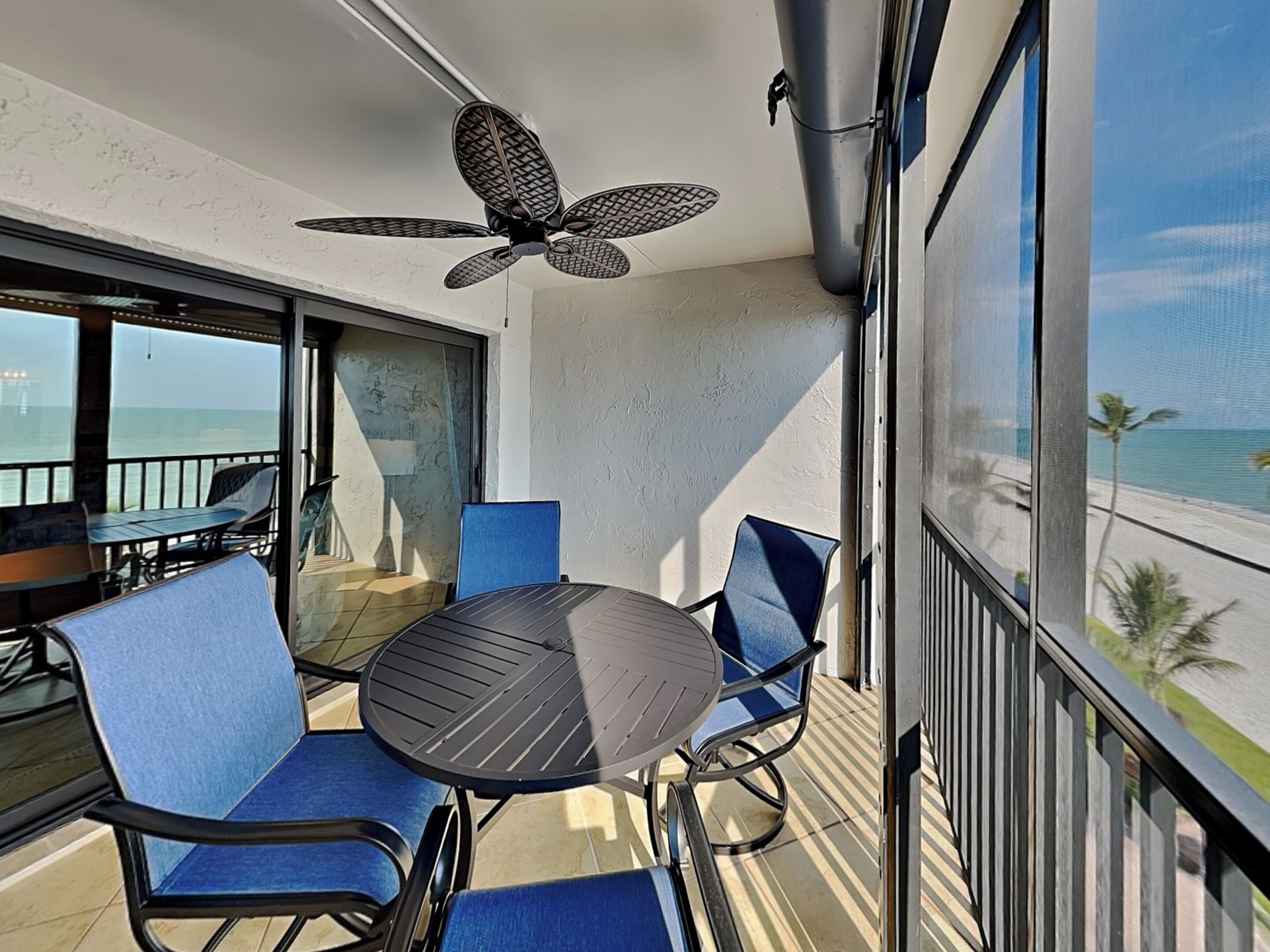 En balkon med et rundt bord og blå stole, en loftsvifte og udsigt til stranden.