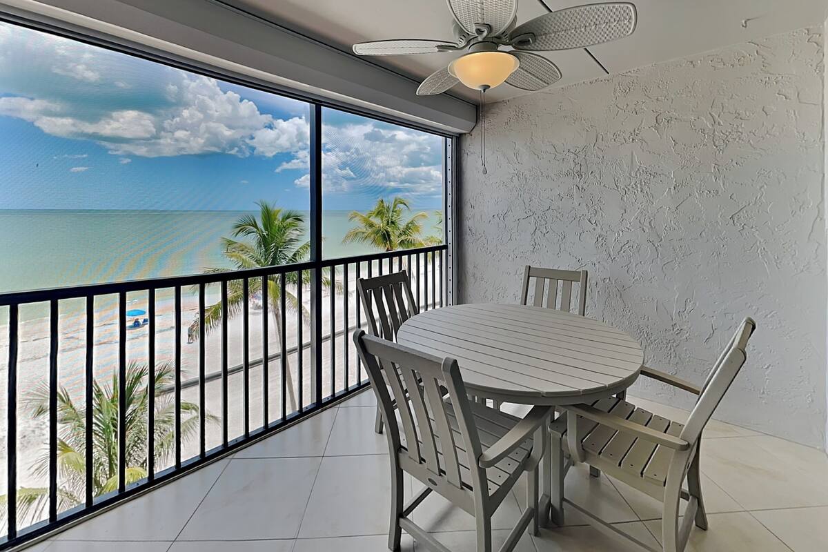 A balcony with a view of the beach and ocean, a round table with chairs, and a ceiling fan.