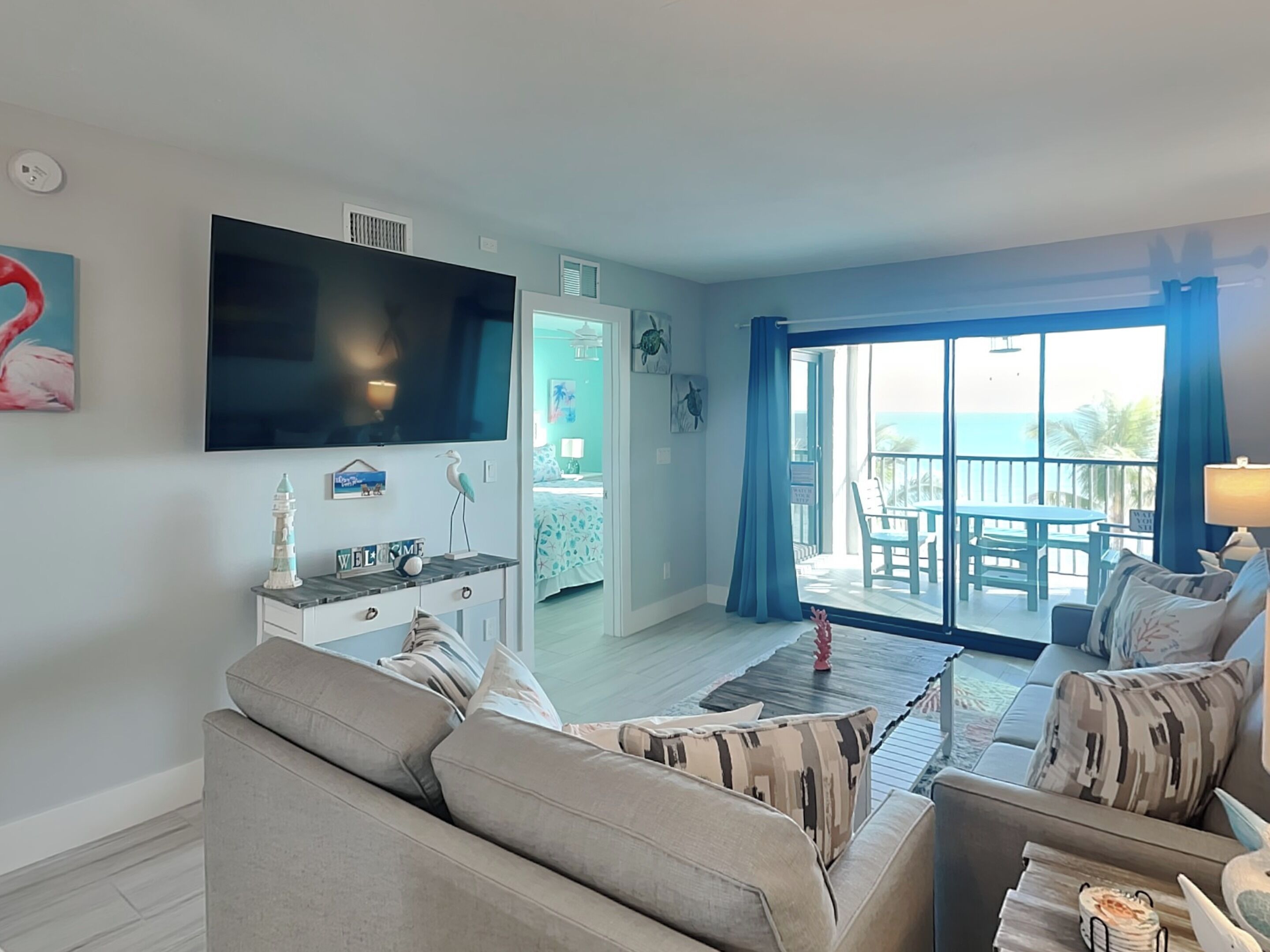 A living room with a flat-screen TV, a sofa, a coffee table, and a balcony with a view of the ocean.
