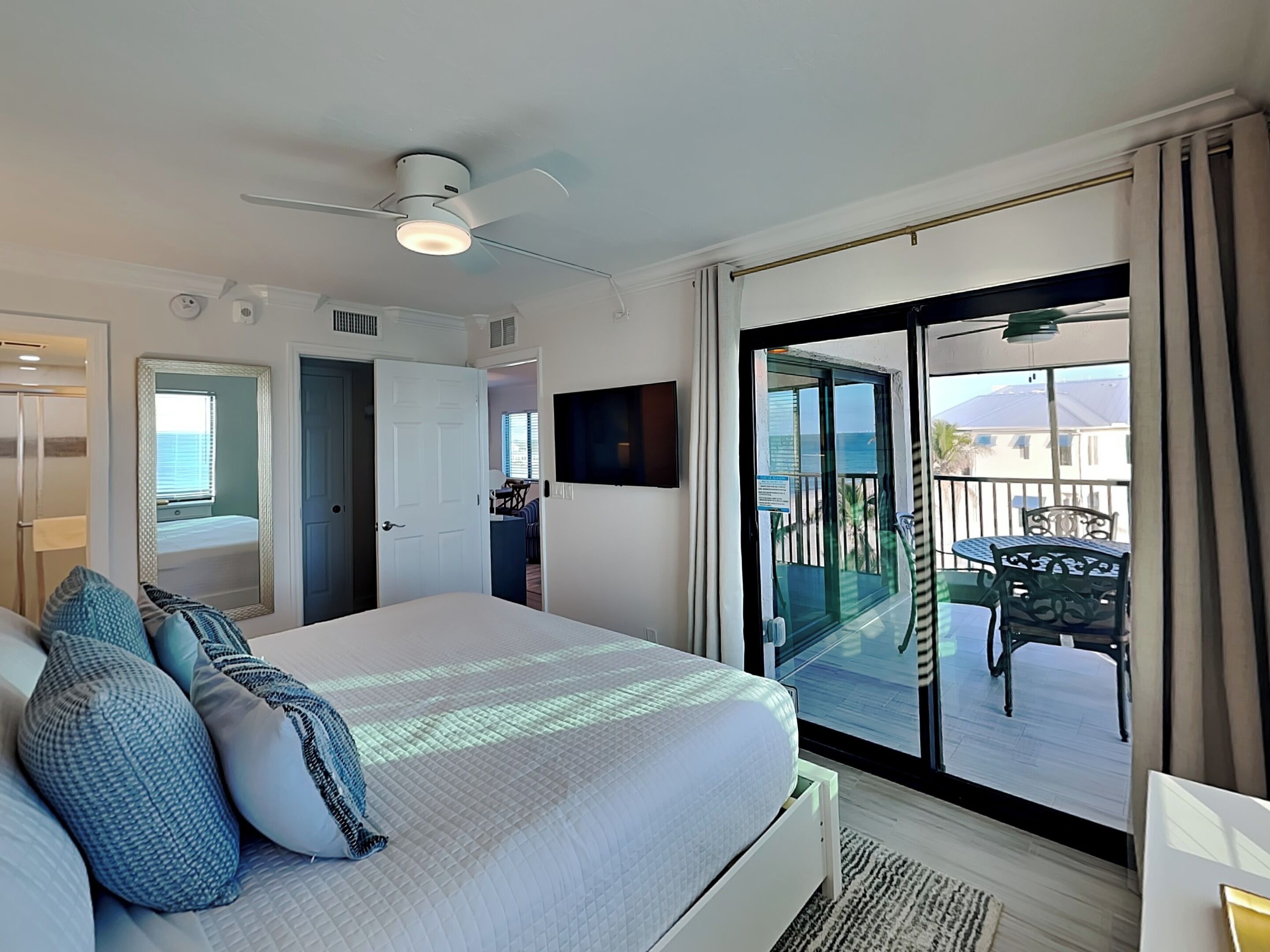 A bedroom with a bed, a ceiling fan, a TV, and a balcony with outdoor furniture.
