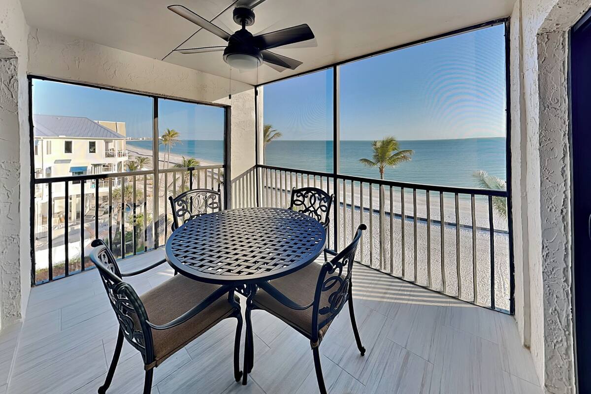 A balcony with a round table and chairs, large windows with a beach view, and a ceiling fan.