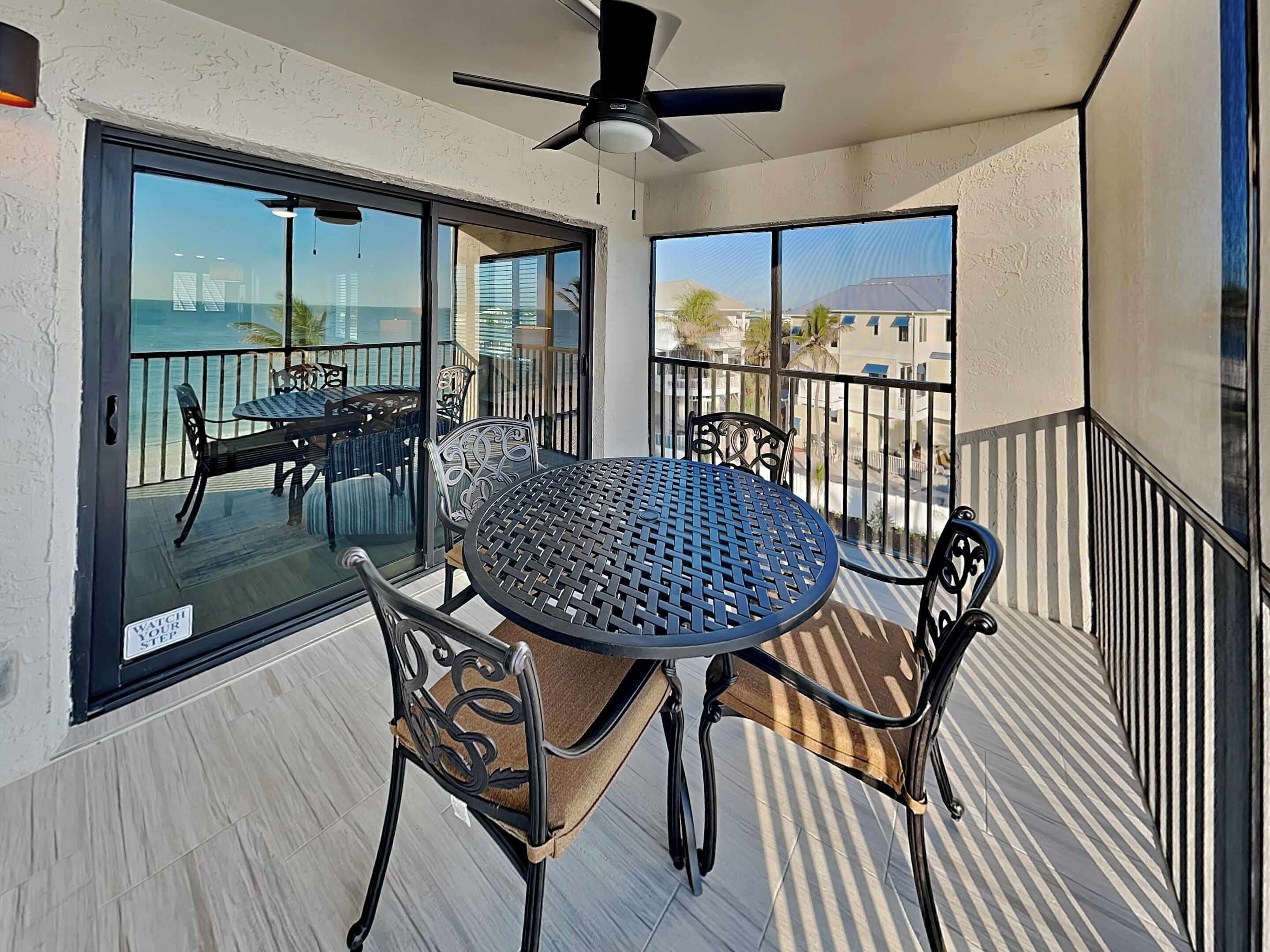 A balcony with a round table and chairs, a ceiling fan, and a view of the ocean and buildings.