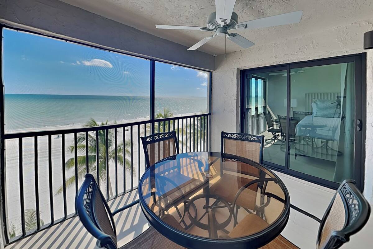 Un balcon avec une table et des chaises en verre, donnant sur une plage et l’océan.
