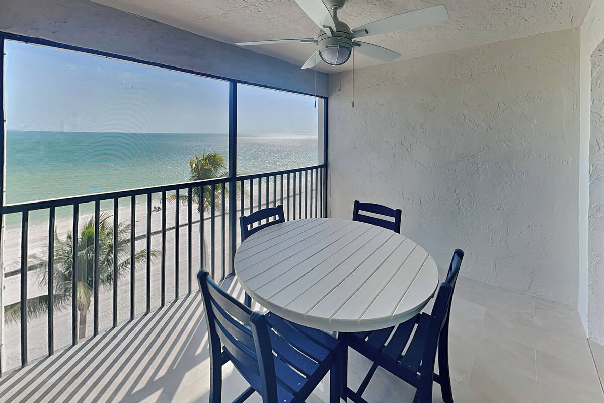 Un balcon avec une table blanche et des chaises bleues, donnant sur une plage et l’océan.