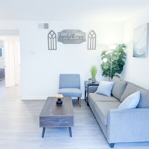 Ein modernes Wohnzimmer mit einem grauen Sofa, einem blauen Sessel und einem Couchtisch aus Holz. An den Wänden befinden sich zwei Dekorationen und eine große Zimmerpflanze.