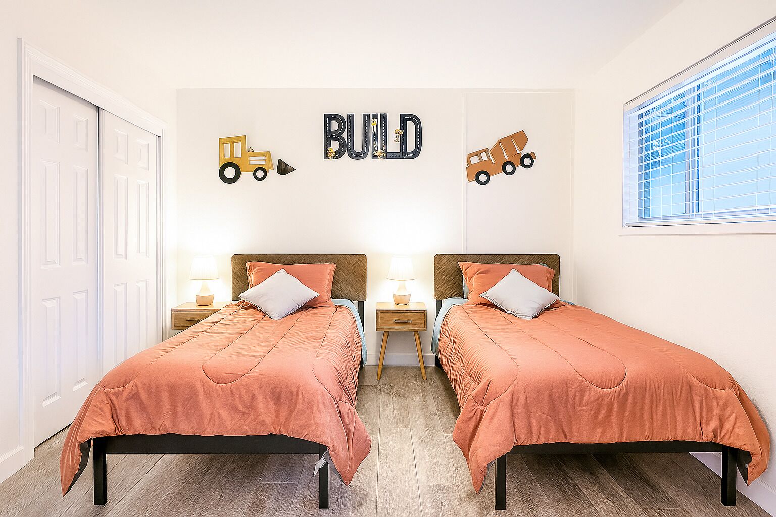 Un dormitorio doble con dos camas, cada una con cabecero, mesitas de noche y lámparas. Las paredes tienen un letrero que dice 'BUILD' y decoraciones con vehículos de construcción.