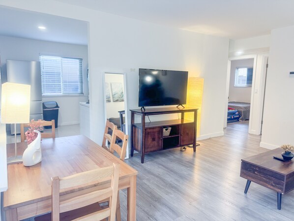 Una sala moderna con una mesa de comedor de madera, sillas, un televisor sobre un mueble bajo y una lámpara.