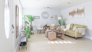 Amplia sala de estar con sofá, sillas, una mesita de centro y una mesa de comedor. Hay plantas en macetas, obras de arte en las paredes y una lámpara en el techo.