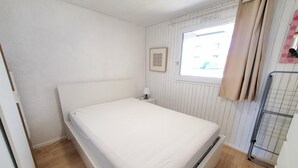 A small, single-bed room with a wooden floor, a white headboard, a window with curtains, and a radiator.