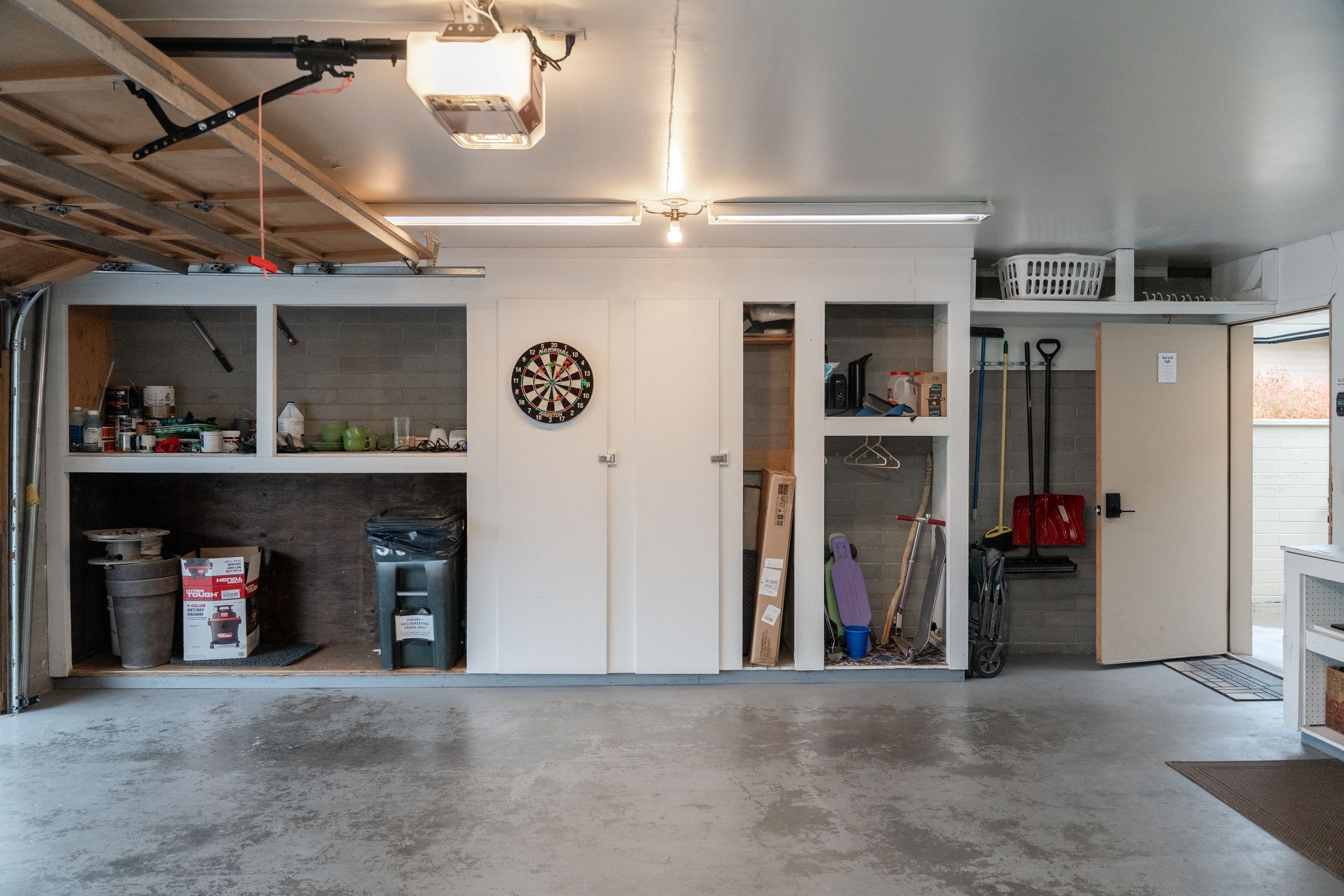 A garage with a concrete floor, white walls, and a ceiling with exposed beams. There are shelves with various items, a dartboard, and a door leading to another room.