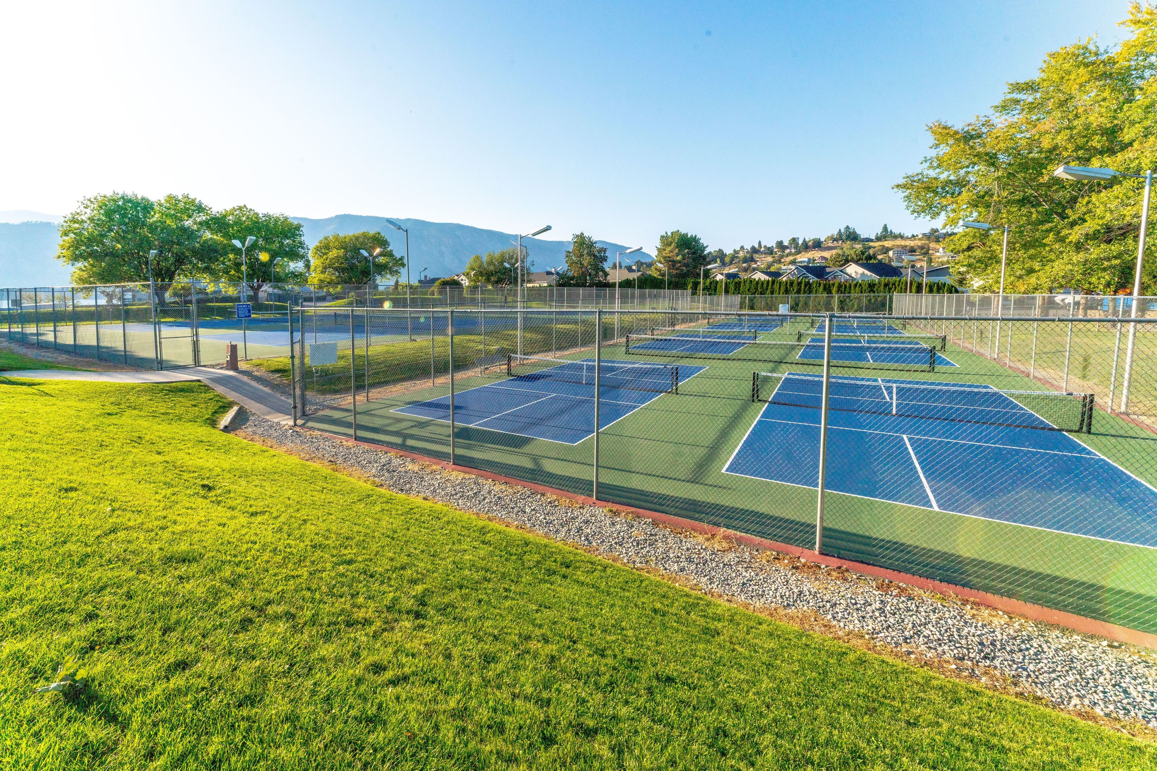 A tennis court with a surrounding fence, a grassy area, and a view of houses and trees in the background.