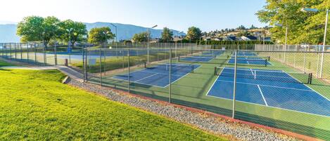 A tennis court with a surrounding fence, a grassy area, and a view of houses and trees in the background.