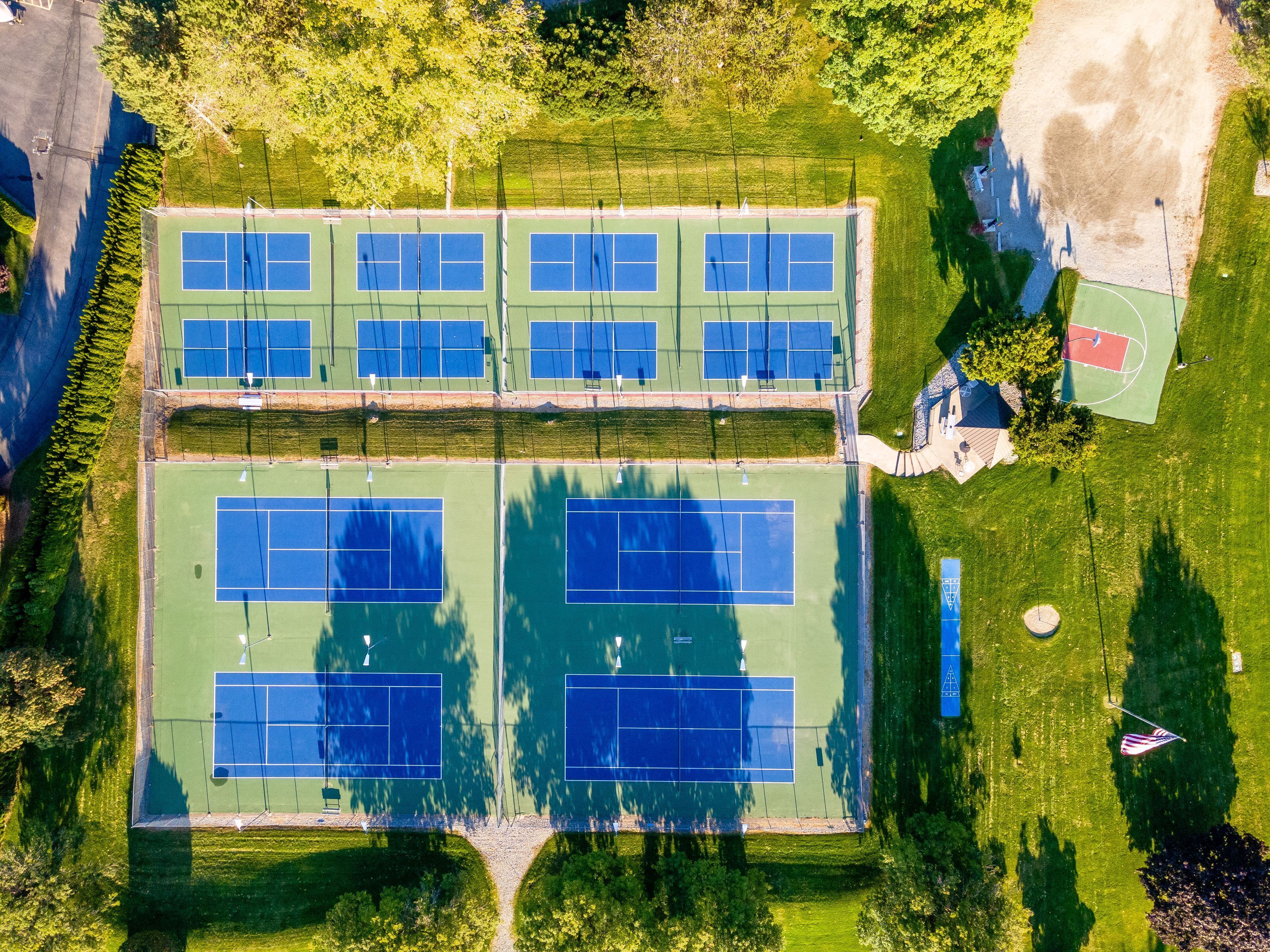 Eine Luftaufnahme eines Sportkomplexes mit mehreren Tennisplätzen, einem Basketballfeld und umgebender Grünfläche.