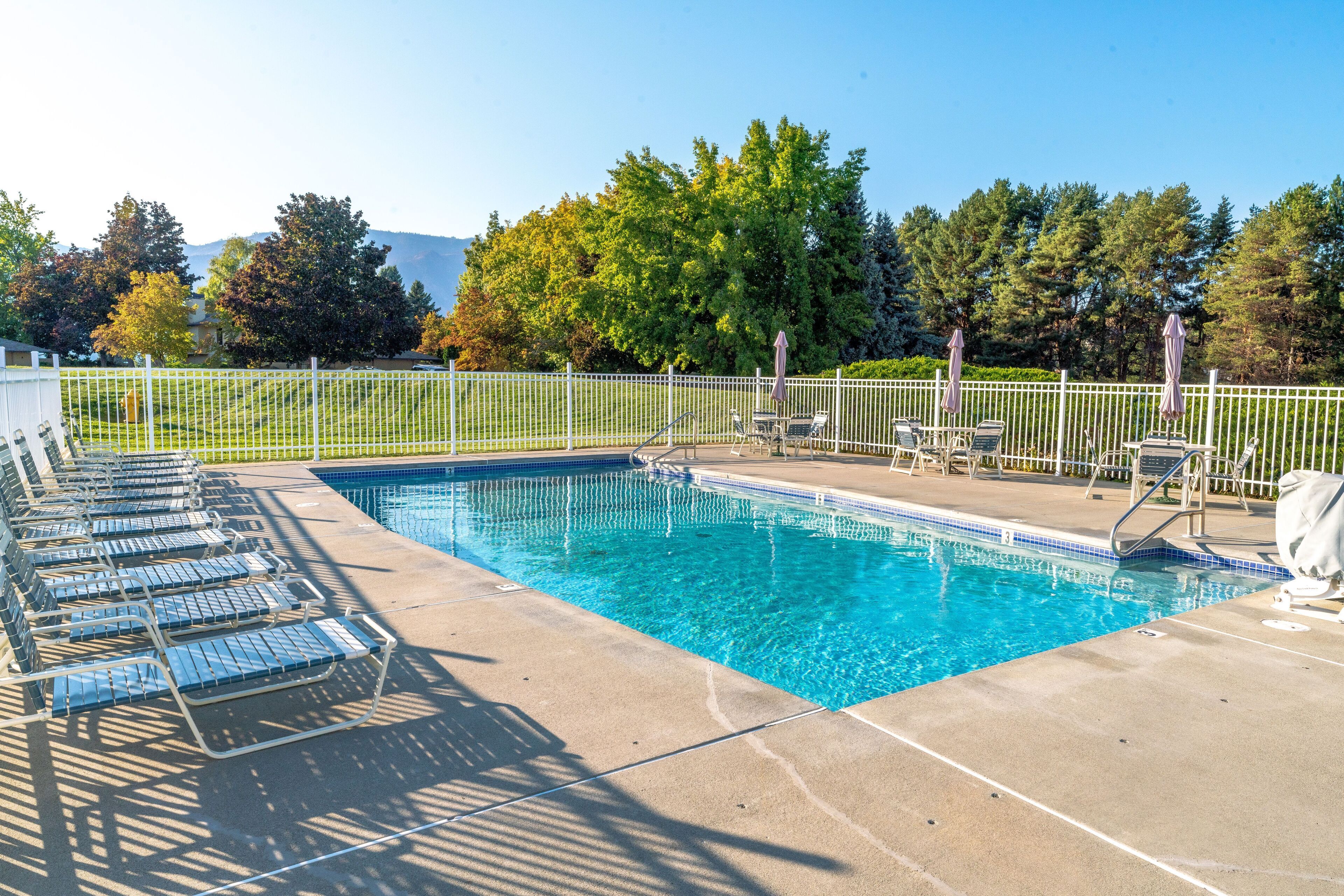 Une piscine rectangulaire entourée d’une clôture blanche et bordée de chaises longues.