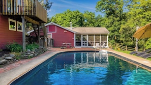 Une piscine avec une terrasse en bois, un bâtiment rouge et un espace couvert avec des chaises.