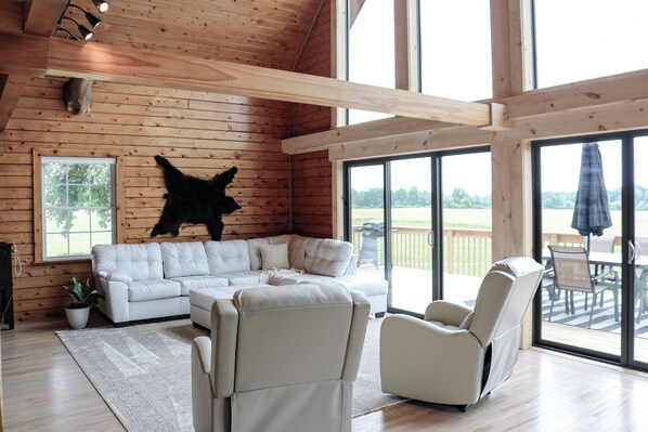 A spacious living room with a white leather sofa, a white armchair, and a large window offering a view of the outdoors. - 3 bedroom secluded Lodge, close to Canandaigua (Canandaigua)