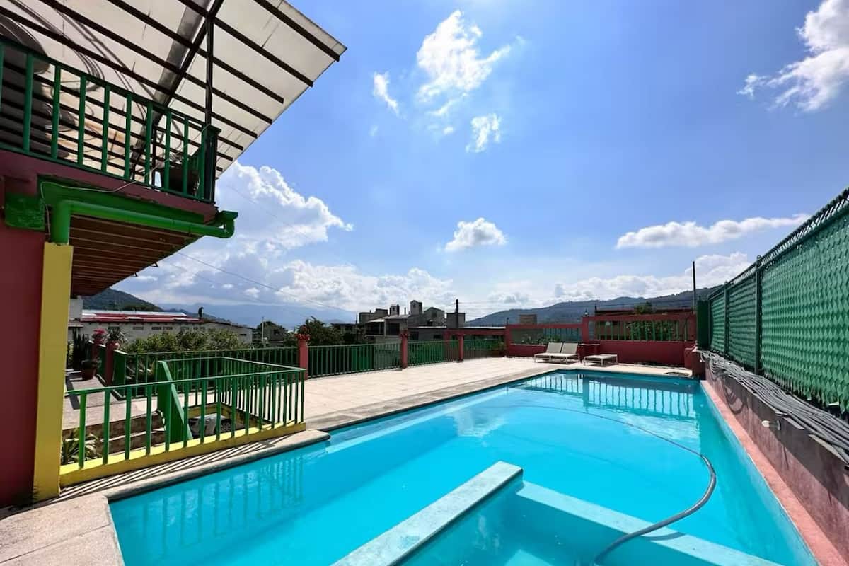Une piscine entourée d’une clôture verte, un espace abrité et une vue sur des bâtiments et des montagnes en arrière-plan.