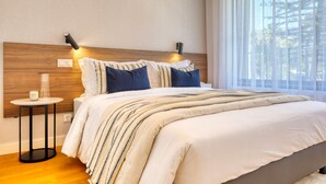 A modern bedroom with a wooden headboard, a bedside table with a lamp, and a large window with sheer curtains.