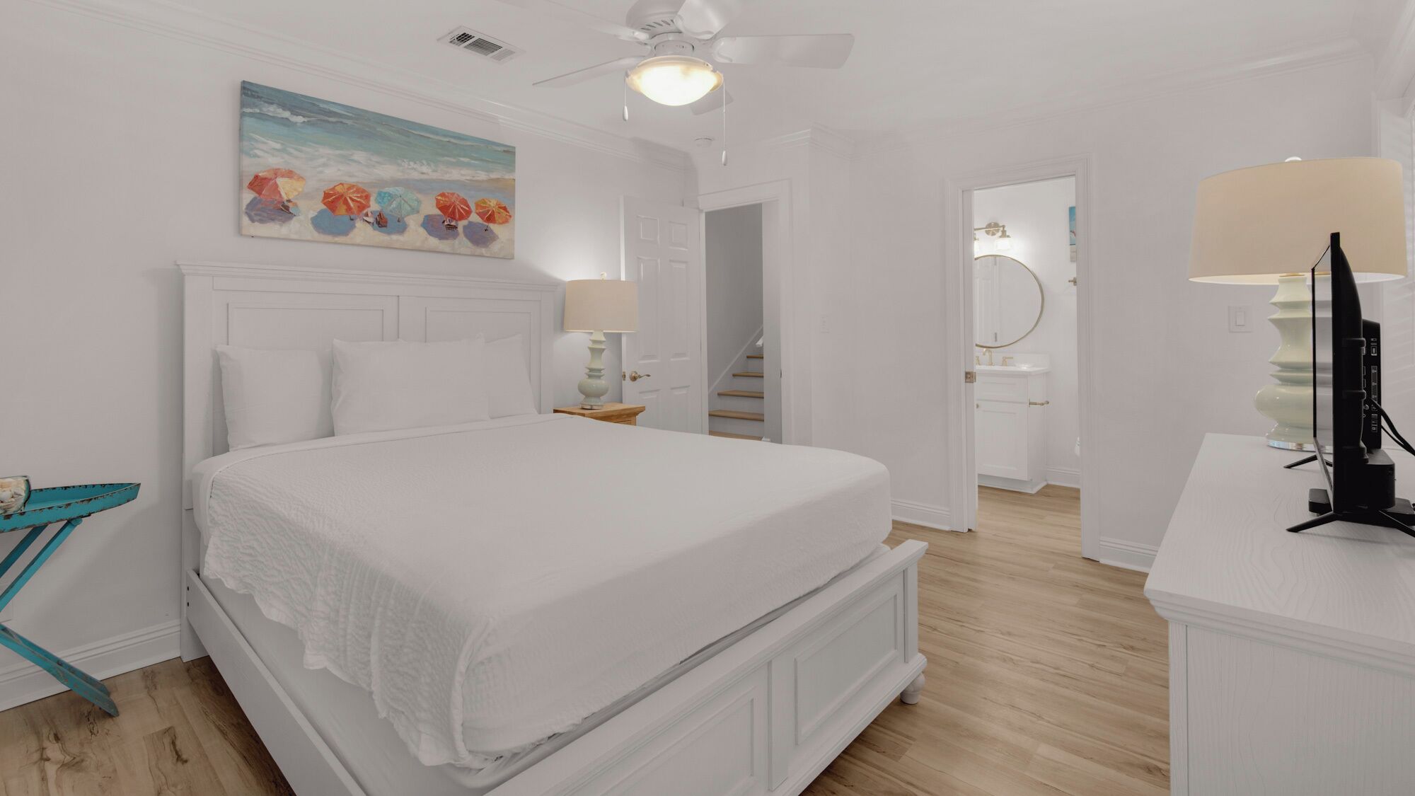 A bedroom with a large bed, a ceiling fan, a painting of beach umbrellas, and a bathroom visible through an open door.