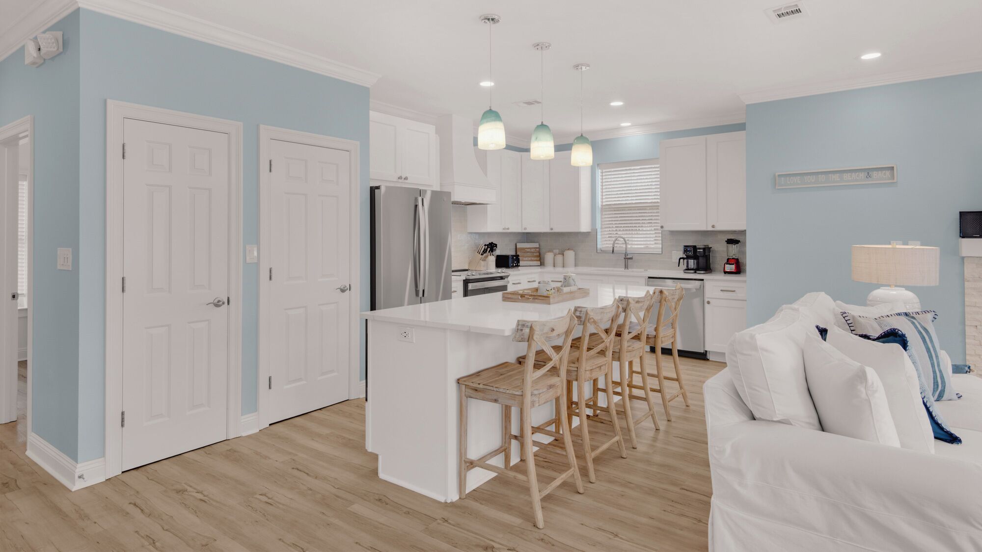 A modern kitchen with white cabinets, a central island with bar stools, and a dining area with a white sofa and cushions.
