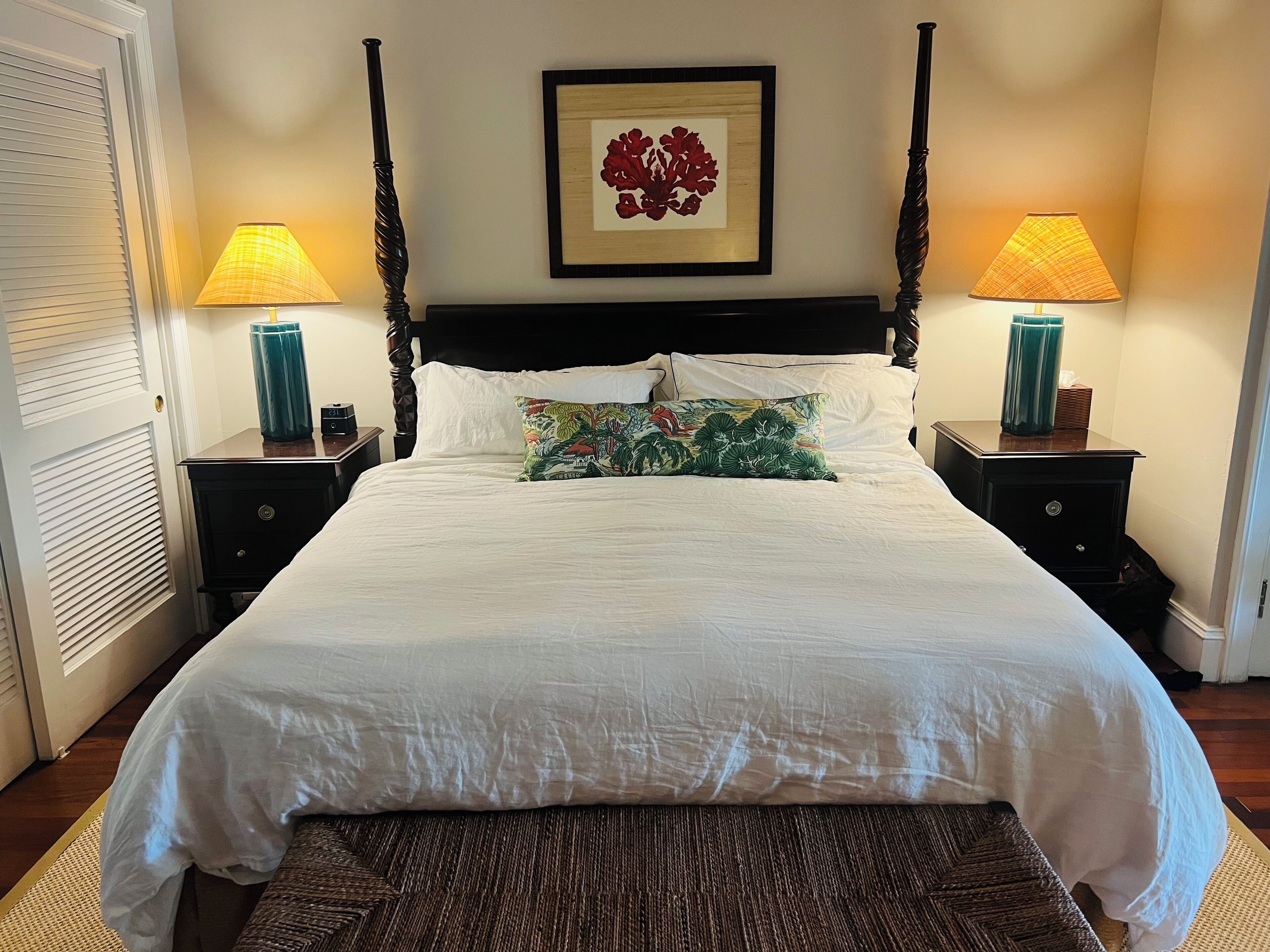 A neatly made bed with white linens, two bedside lamps, a framed artwork above the bed, and a wooden floor.