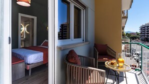 Un balcón con una cama, una silla de mimbre, una mesita con desayuno y vistas a la calle y edificios.