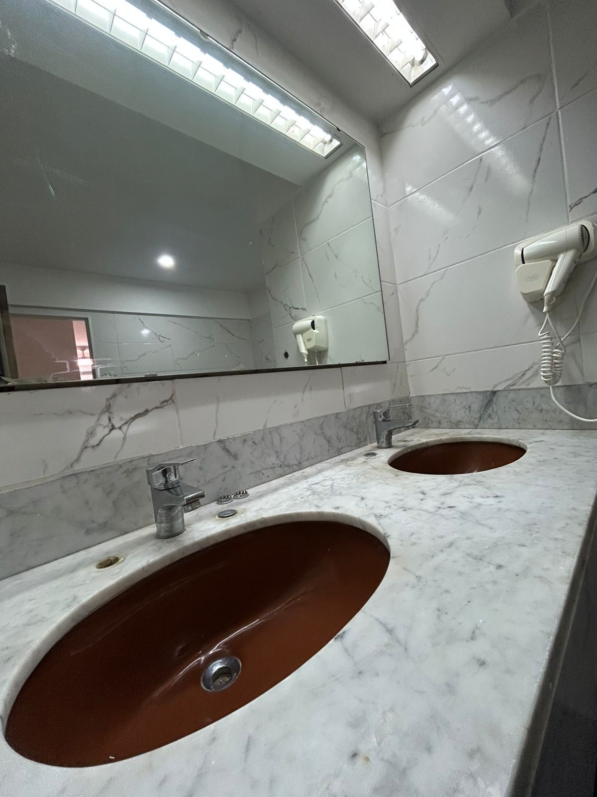 Un baño con dos lavamanos, un espejo y un secador de pelo fijado a la pared.