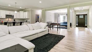 Ein modernes Wohnzimmer mit einer Eckcouch, einem Essbereich mit Tisch und Stühlen sowie einer Küche mit weißen Schränken und einer Mikrowelle.