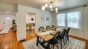 A dining area with a wooden table set for six, black chairs, a chandelier, and a view of the outdoors through large windows. - 7 Mi to Mt Rainier NP: Home w/ Private Hot Tub! (Ashford)