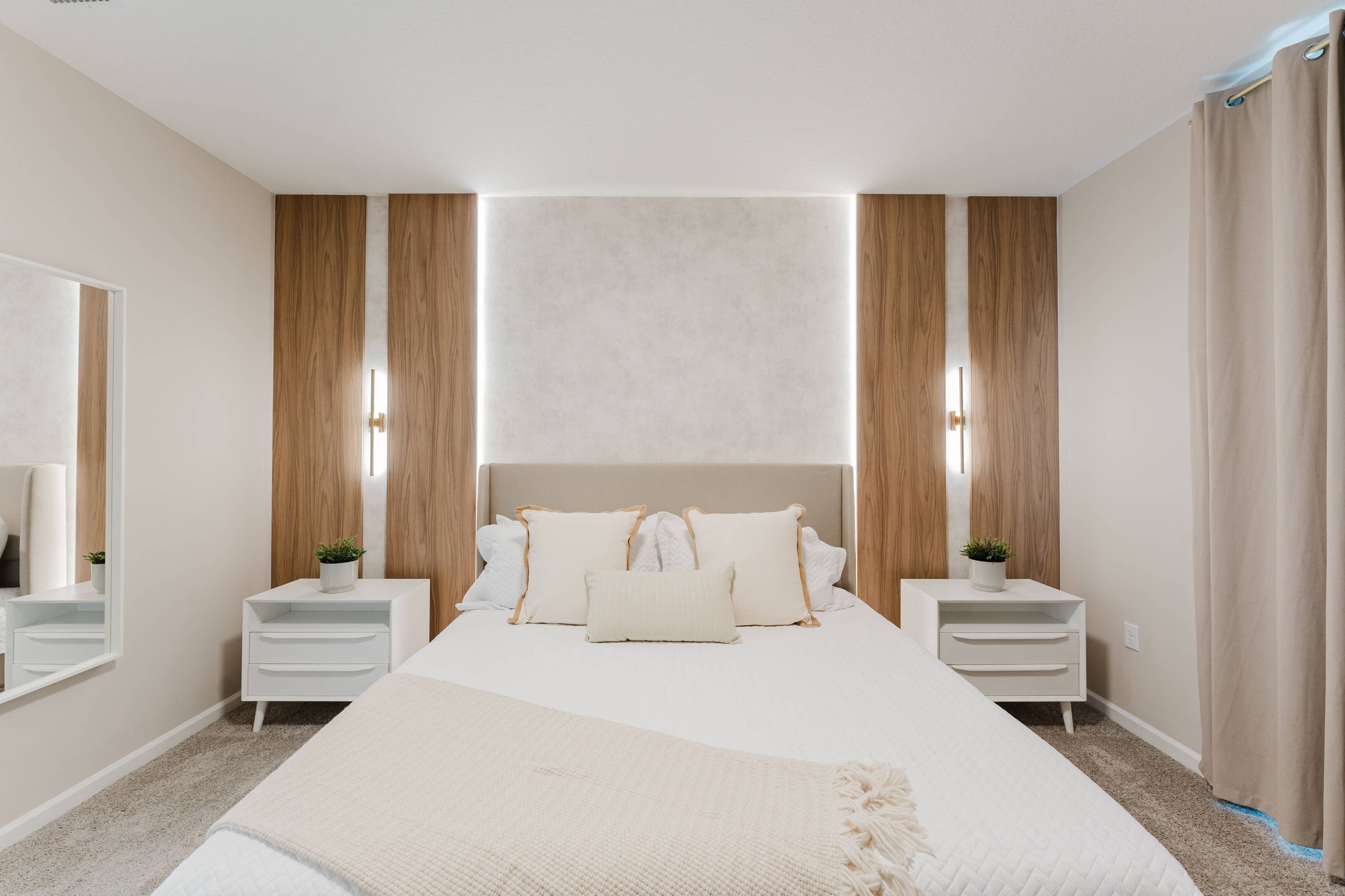 A modern bedroom with a large bed, two bedside tables, a mirror, and a headboard with built-in lighting.
