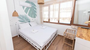 A bedroom with a bed, a wooden chair, a window with lace curtains, and a wall-mounted air conditioner.