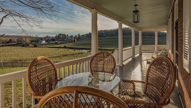 Une véranda couverte, dotée de fauteuils en osier et d’une table en verre, qui offre une vue sur un paysage rural.