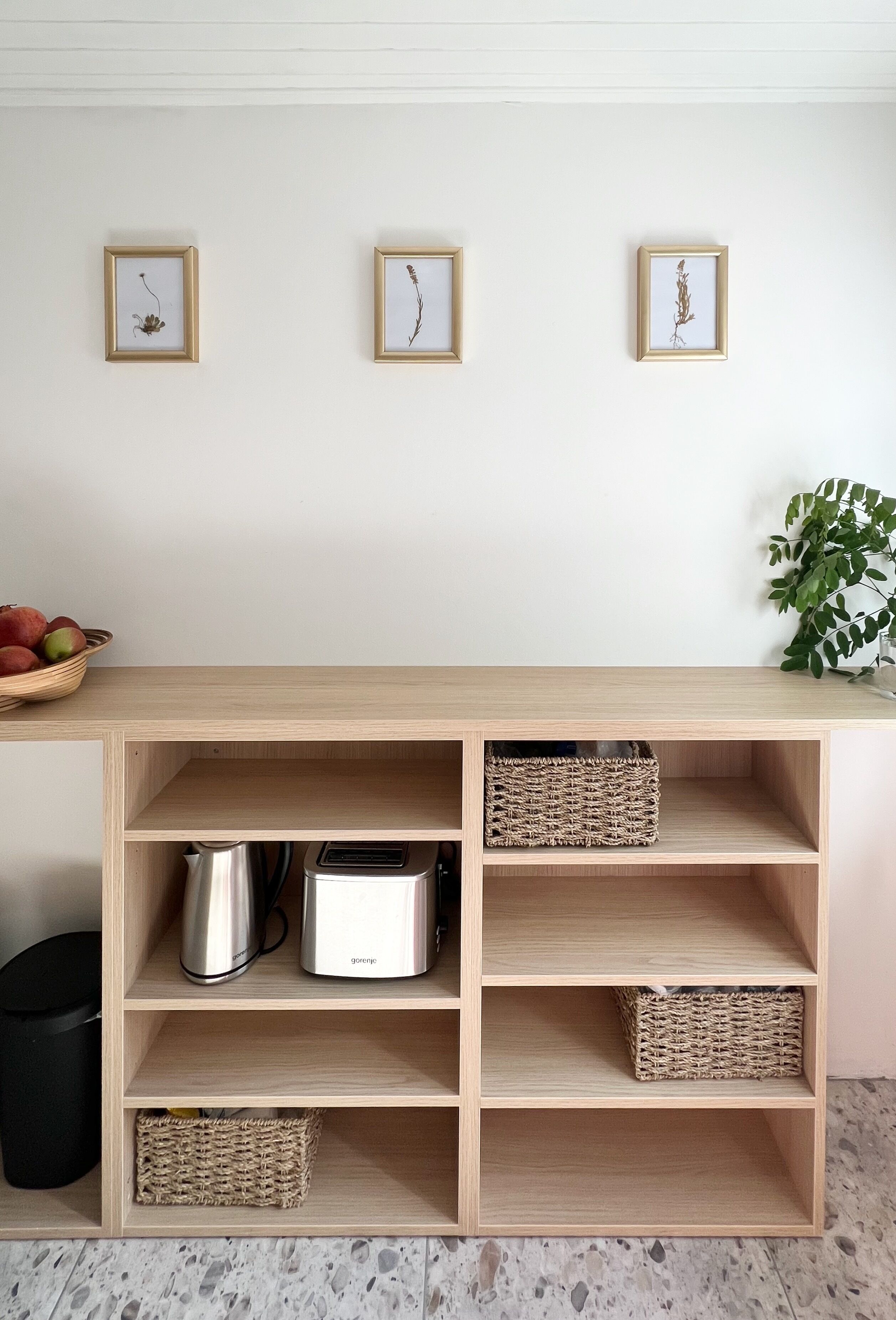 A kitchen with wooden shelves, a toaster, a kettle, and three framed pictures on the wall.