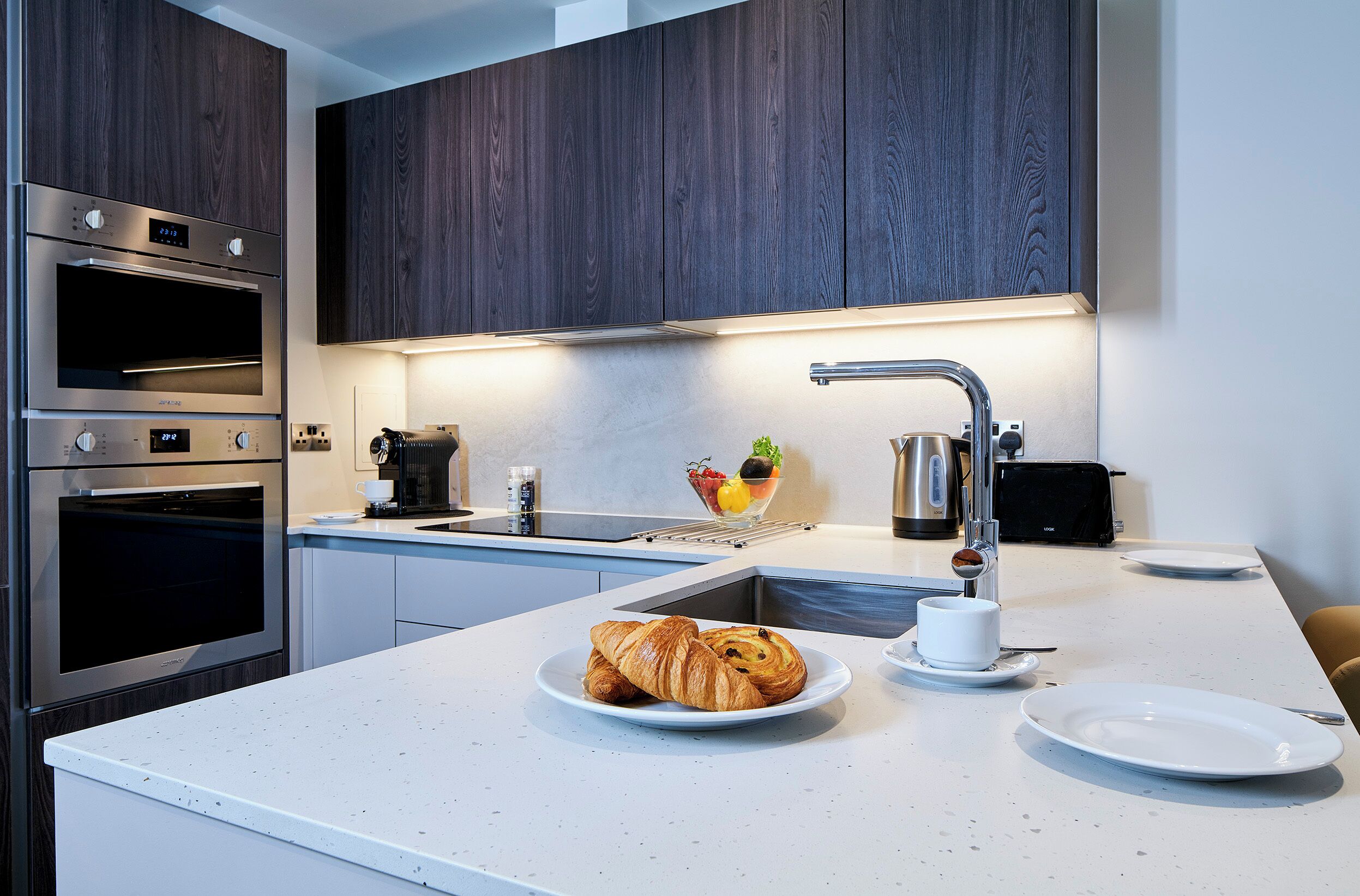 Et moderne køkken med indbyggede ovne, en stålvask og et morgenmåltidsområde med croissanter og en kaffekop.