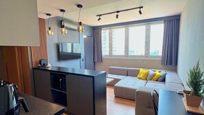 Un apartamento moderno con cocina, comedor y sala de estar. Cuenta con un ventanal grande, un televisor de pantalla plana y una pequeña mesa de comedor.
