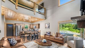 A spacious living area with a high ceiling, wooden beams, a kitchen with modern appliances, and a view of greenery outside.